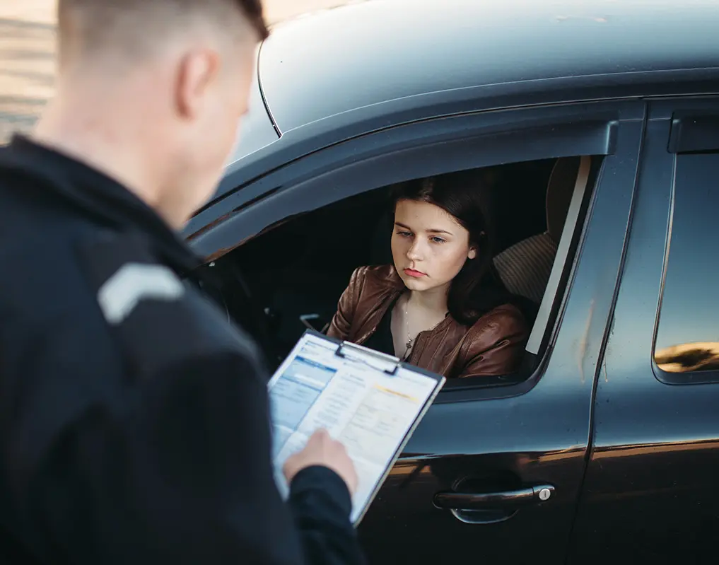 A person in a car looks at an officer standing outside the vehicle holding a clipboard.