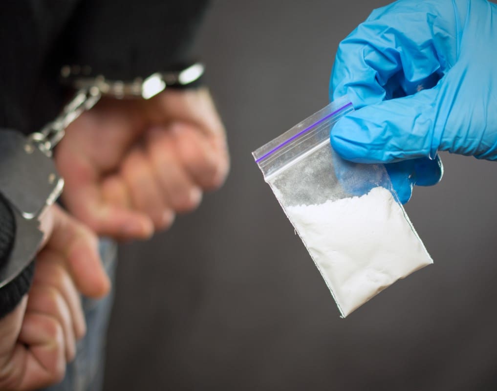 A person in handcuffs is shown next to a gloved hand holding a small plastic bag of white powder.