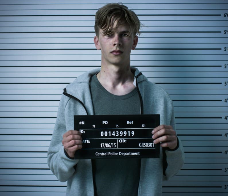 A young man stands against a height chart holding a police identification board.