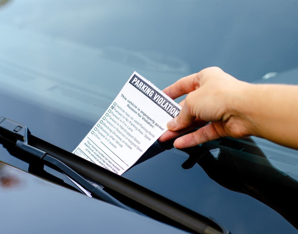 A hand places a parking violation ticket under a car's windshield wiper.