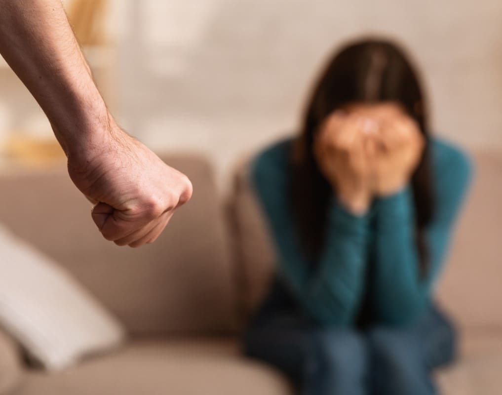 A person's clenched fist in the foreground, while a woman sits on a couch in the background, covering her face with her hands, suggesting a tense or distressing situation.