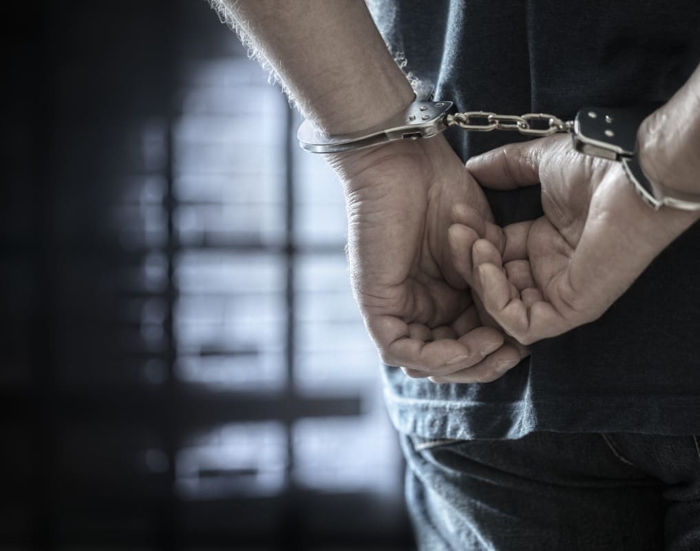 A person in handcuffs stands with hands behind their back in a dimly lit room with barred windows.