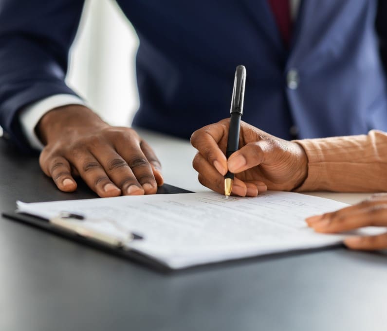 Two people at a desk, with one signing a document while the other watches closely, emphasizing collaboration or agreement.