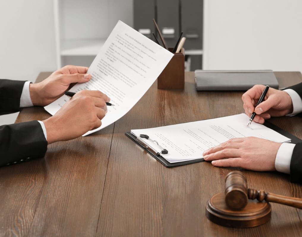 Two people in suits are seated at a table, examining legal documents. One person holds a pen over a clipboard, while the other holds a paper. A gavel is placed on the table.