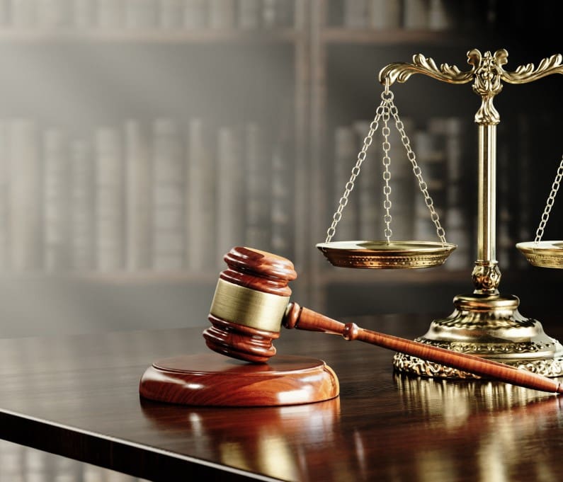 A wooden gavel and bronze scales of justice on a polished desk, with bookshelves in the background.