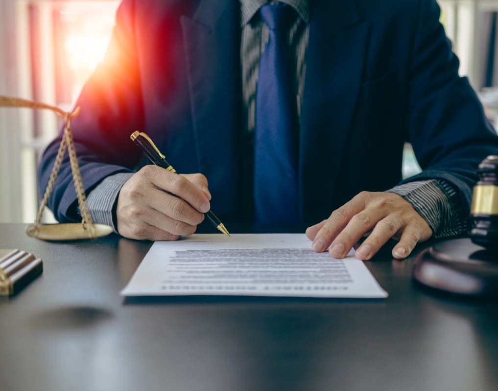 A person in a suit signs a document at a desk with a gavel and scales of justice nearby.