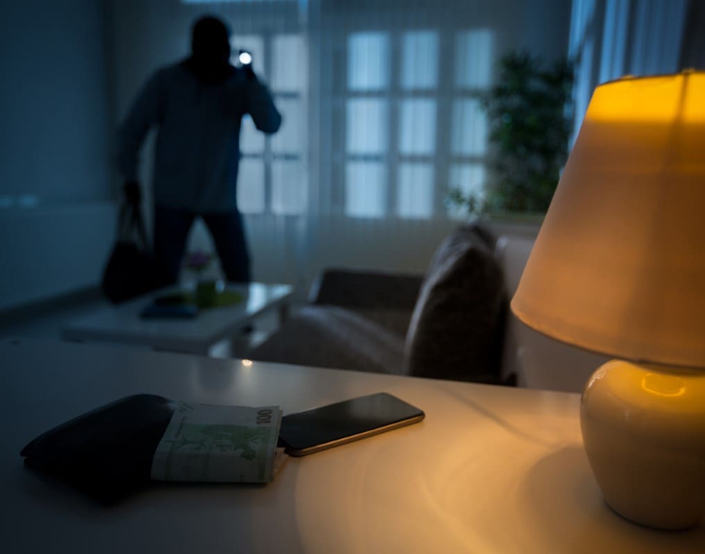 Dimly lit room with a lamp, a wallet, and a phone on a table. A shadowy figure stands in the background, holding a bag.