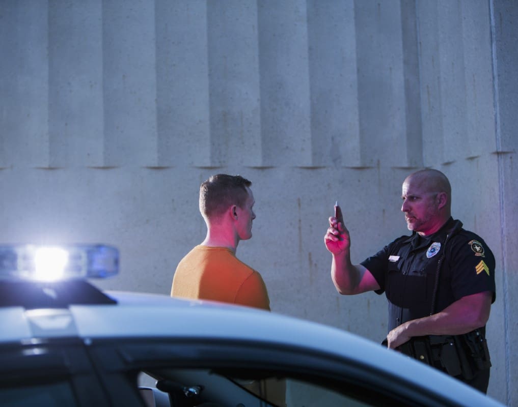 Police officer conducts a sobriety test on a man, holding up a finger. A police car with flashing lights is in the foreground.