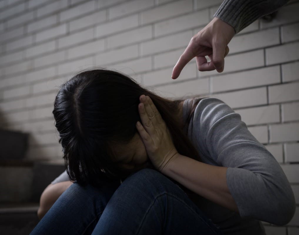 Person crouched on stairs, covering ears, while another person's hand points at them in an accusatory manner.