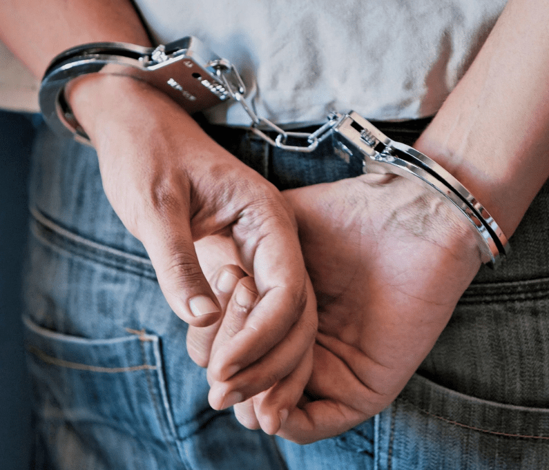 Person wearing handcuffs stands with hands behind back, wearing casual jeans and a white shirt.