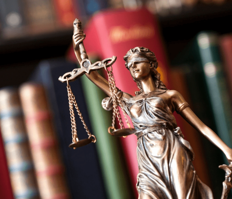 Close-up of a Lady Justice statue holding scales, with colorful books in the blurred background.