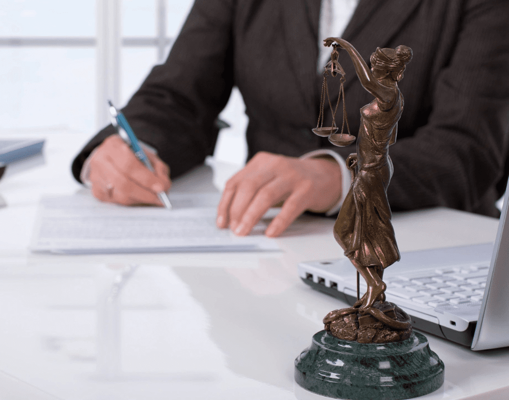 A bronze statue of Lady Justice on a desk next to a laptop. A person in a suit is writing on a document in the background.